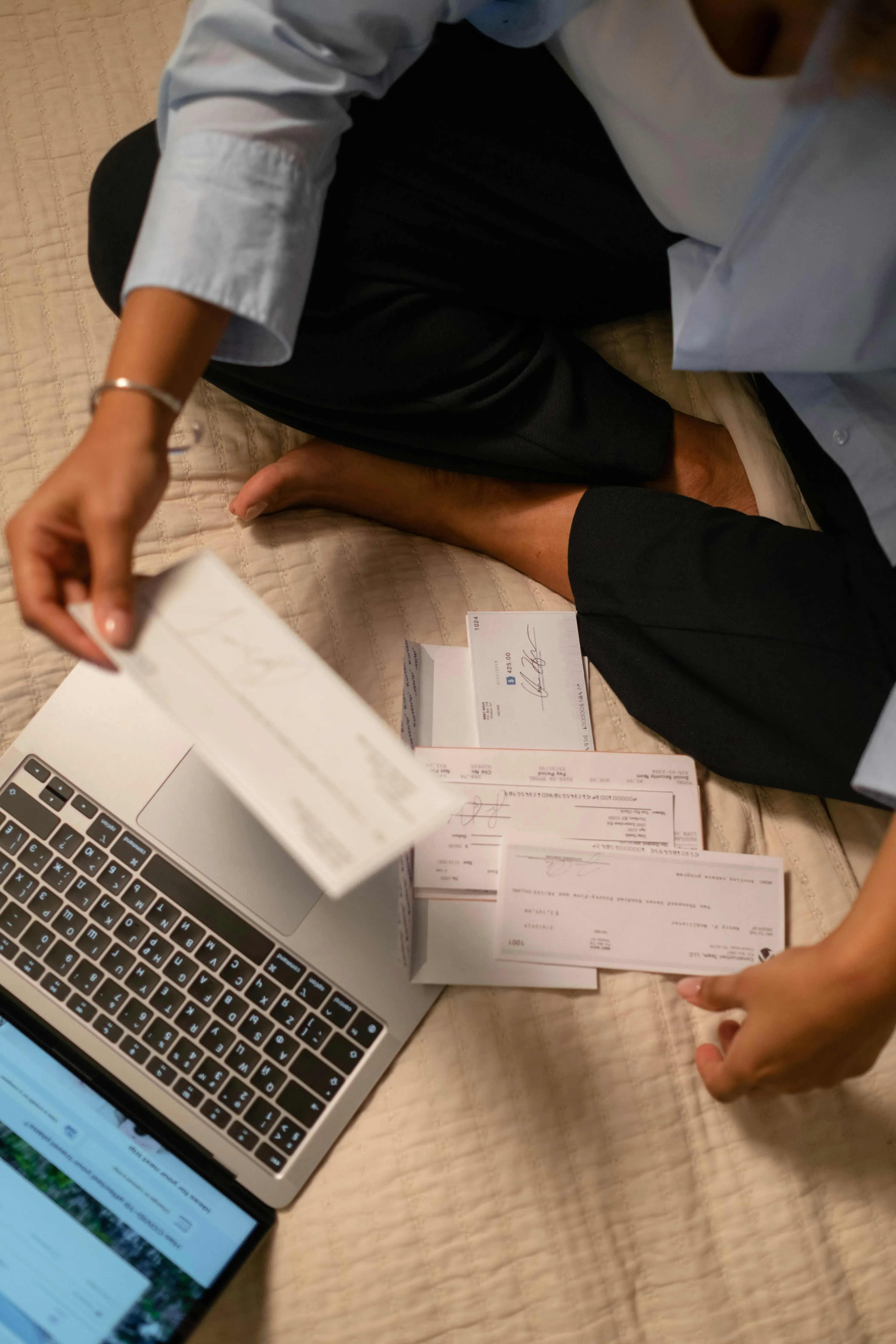 Picture of a person sitting on a bed with a laptop while reviewing printed paper documents